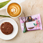 Byron Bay Cookies Triple Choc Gluten Free Cookie unwrapped on white plate bottom left, wrapped cookie bottom right, pictured with coffee top left on a sandstone benchtop with green leaves top left corner.