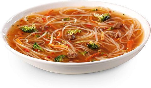 White china bowl filled with a transparent beef broth containing carrot, broccoli, capsicum, rice noodles and pieces of beef meat.