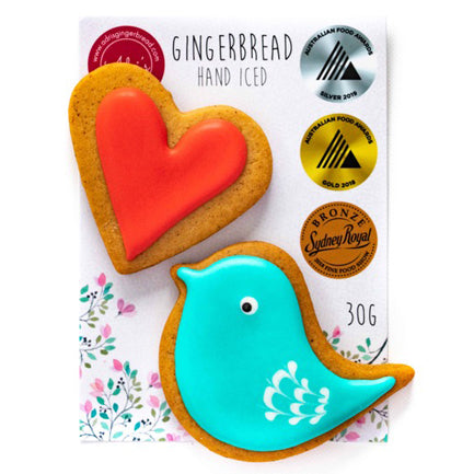 Hand iced blue bird gluten free gingerbread biscuit and red heart gluten free biscuit on top of white packaging card with Adri's Gingerbread logo in top left corner, three food award medal icons down right side and flower motif along bottom.